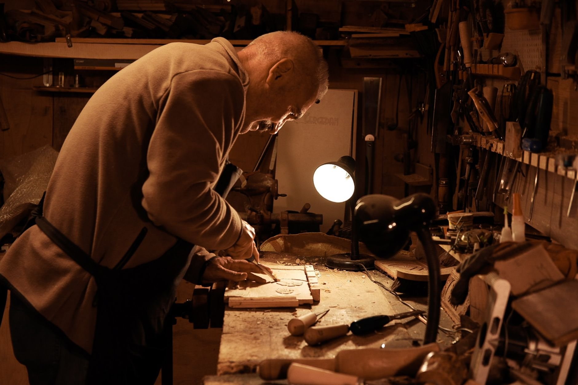 A dedicated senior artisan working on a detailed wood carving in a warmly lit workshop.